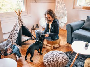 Woman sitting on a stool playing with her small black dog. They are in the living room, there is a dog teepee. 